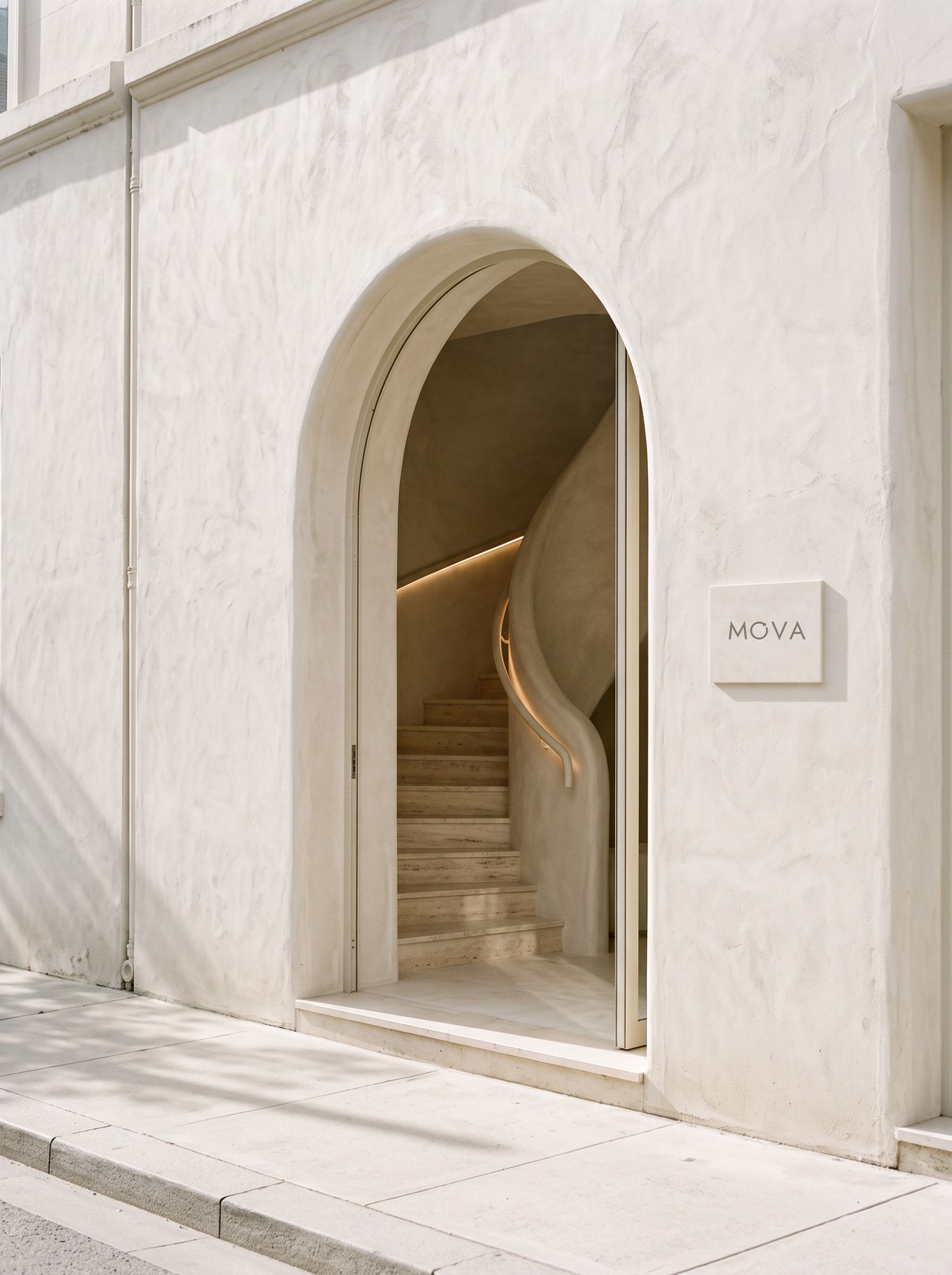 An arched street doorway opening onto a curved limewash stair with travertine treads, lit along the handrail.