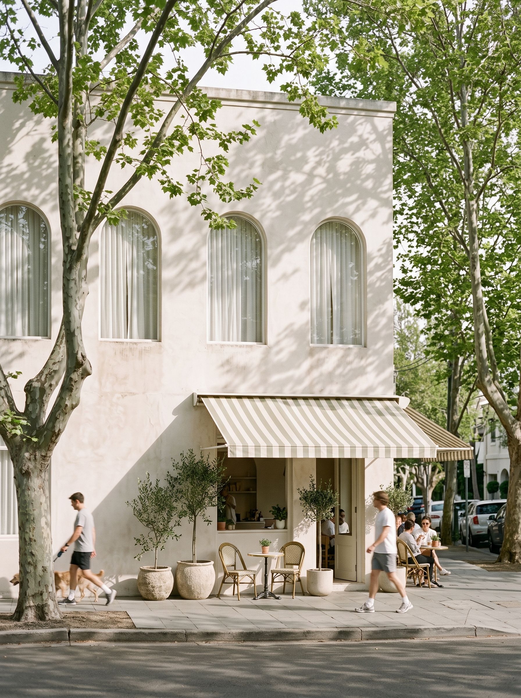 A leafy harbourside village street in early morning, a pale building with arched first-floor windows above a café awning.