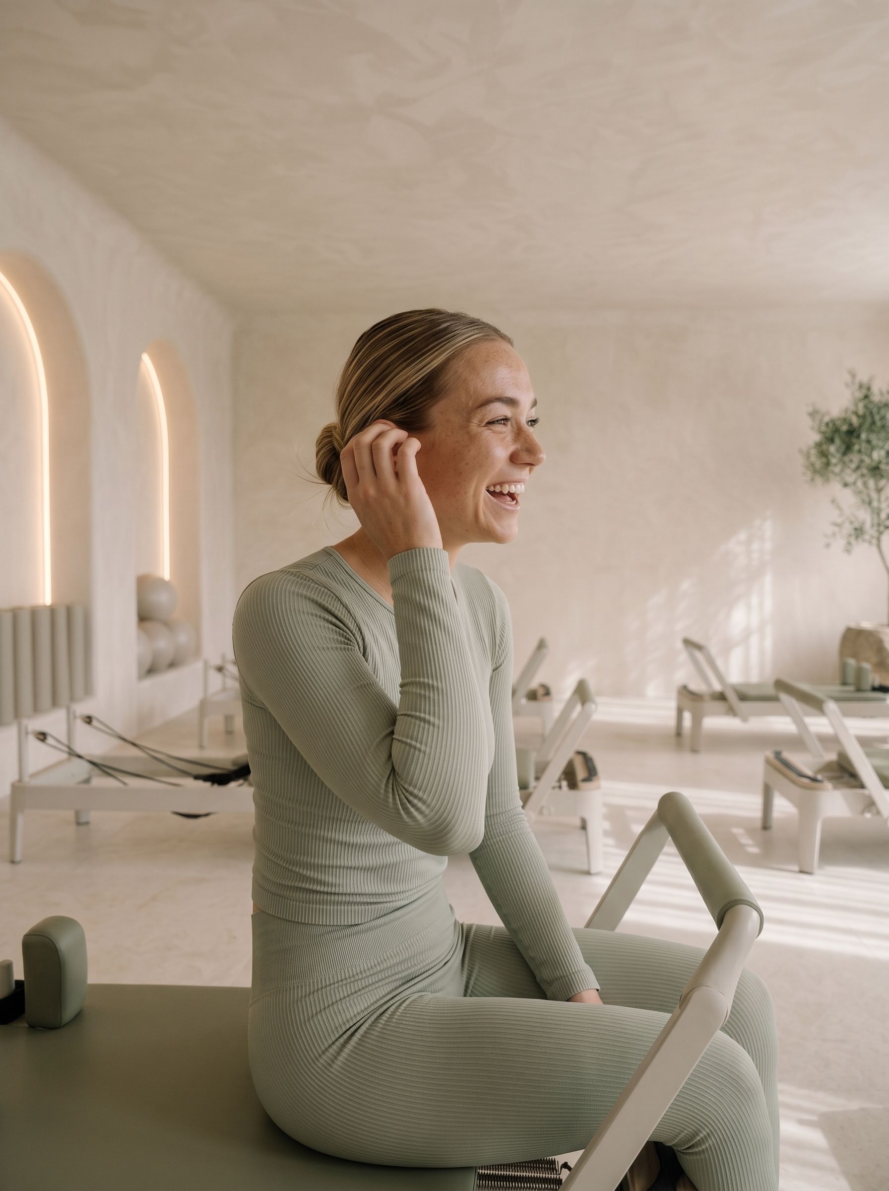 Georgia Hale sitting sideways on a reformer, laughing with someone out of frame.